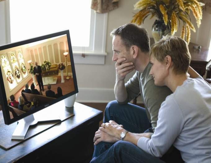 A couple holding hands watching a funeral service on a computer in a living room 