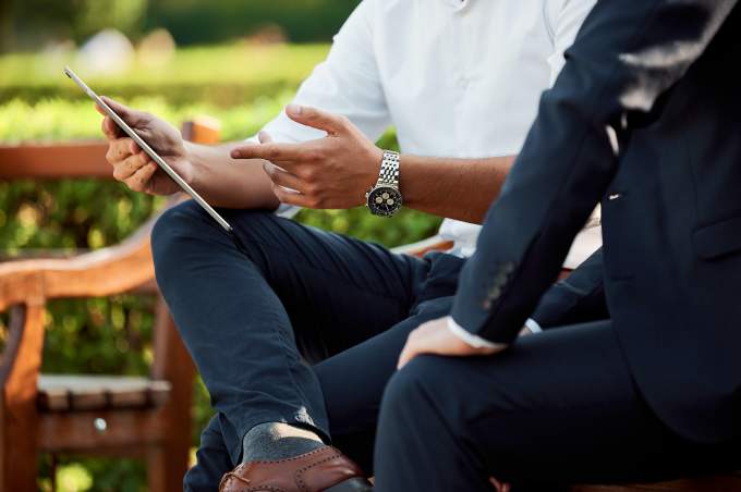 A funeral director on his tablet talking to another funeral director on a garden bench outside a funeral home 